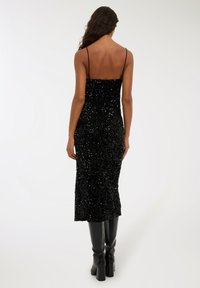 Kobieta z długimi włosami, ubrana w czarną cekinową sukienkę midi na cienkich ramiączkach i czarne wysokie botki, stoi tyłem.
