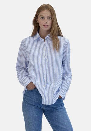 Donna con lunghi capelli rossi che indossa una camicia azzurra a righe con bottoni e jeans blu, in piedi con le mani in tasca davanti a uno sfondo bianco.
