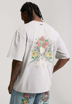 Uomo con capelli intrecciati che indossa una maglietta grigio chiaro con un teschio dalle ali di farfalla e rose colorate sulla schiena, abbinata a pantaloncini di jeans ricamati.