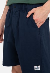 Navy blue shorts with an elastic waistband, side pockets, and a logo patch featuring a mountain design at the hem. Smooth fabric texture.