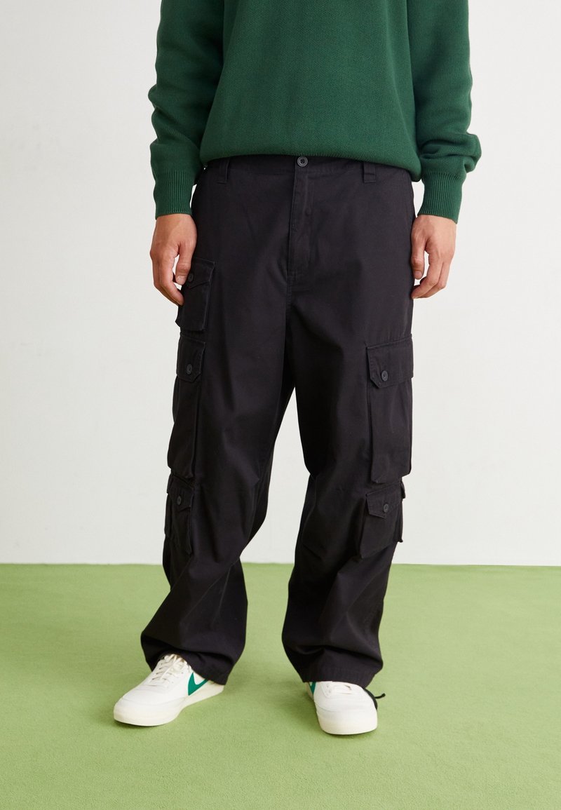 Schwarze Cargo-Hose mit mehreren Seitentaschen, entspanntem Schnitt und Knopfverschluss. Kombiniert mit weißen Sneakers mit grünen Akzenten.