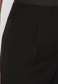 Pantalon noir avec une texture lisse, doté de plis marqués, de poches latérales et d'un design ajusté. Le matériau présente un léger brillant.