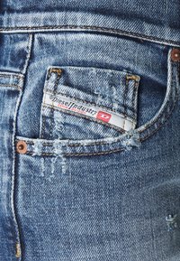 Denim jeans with a faded blue wash, featuring distressed elements and a small front pocket displaying a red and white label.