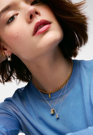 Kobieta z krótkimi brązowymi włosami, ubrana w niebieską bluzkę, złoty naszyjnik z koralików, dwa srebrne naszyjniki z zawieszkami oraz złote i srebrne kolczyki.