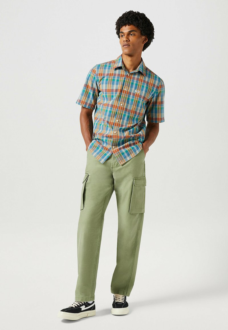 Camisa de cuadros de manga corta en azul, marrón y verde, combinada con pantalones cargo en oliva claro con bolsillos laterales y zapatillas negras.