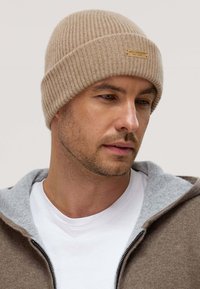 Beige ribbed knit beanie made of soft material, featuring a brown label with embossed text. Worn with a gray hooded jacket over a white shirt.