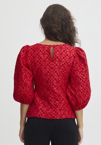 Blouse rouge texturée avec un motif floral, des manches bouffantes et un détail en forme de clé à l'arrière. Le matériau semble léger et tissé.
