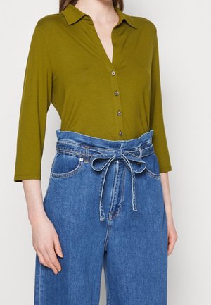 Femme portant une chemise boutonnée vert olive à manches trois-quarts et un jean bleu taille haute noué avec une ceinture en tissu en nœud.