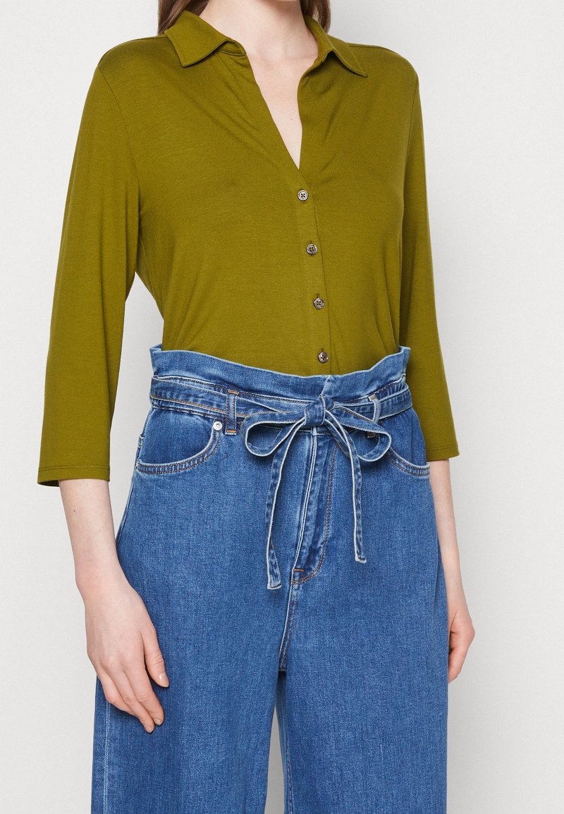 Femme portant une chemise boutonnée vert olive à manches trois-quarts et un jean bleu taille haute noué avec une ceinture en tissu en nœud.