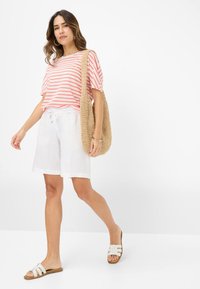 Stribet pink og hvid kortærmet top, hvide linned shorts og brune slidesandaler. Bærer en vævet beige taske, står på en hvid baggrund.