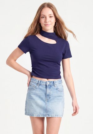 Jeune femme aux longs cheveux portant un crop top bleu marine avec découpe et une jupe en jean bleu clair, debout avec une main sur la hanche.
