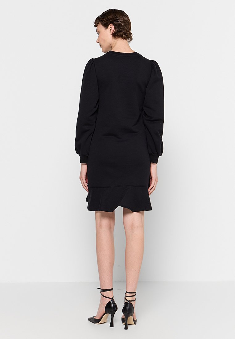 Robe noire en tissu doux, avec des manches longues bouffantes, un col rond et un ourlet volanté. Associée à des chaussures à talons hauts noires.