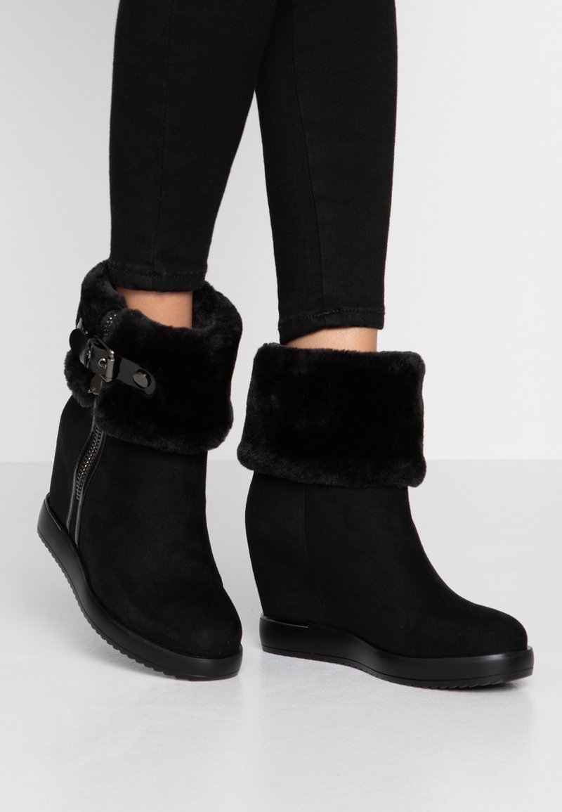 Bottes compensées noires à la cheville avec poignets en fausse fourrure rabattus et fermeture éclair latérale, portées avec un pantalon noir.