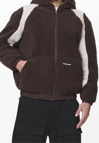 Giacca in fleece marrone con una patch color crema, chiusura a zip frontale e cappuccio. Vita e polsini elasticizzati, con un logo sulla tasca inferiore.
