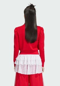 Červený top s dlhými rukávmi, z vypchanými ramenami a priliehavým pásom. Viacvrstvová biela volániková spodná časť dodáva textúru. Vlasy sú upravené do drdolu.
