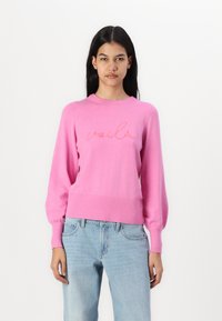 Pull en tricot rose à manches longues, avec un ourlet et des poignets côtelés. Présente "voilà" brodé en couleur orange contrastante sur le devant.