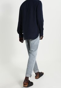 Camicia a maniche lunghe blu navy, jeans chiari tapered e sandali neri con suole in sughero, mostrati da dietro, enfatizzando le trame e la vestibilità dei capi.