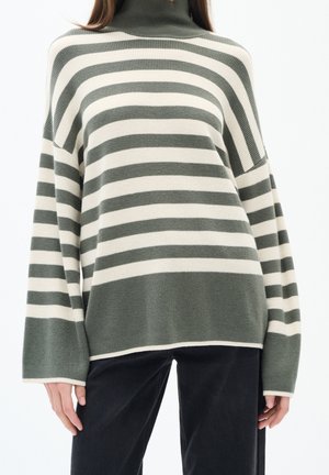 Kvinde iført oversized olivengrøn og hvidstribet sweater med rullekrave og sorte bukser mod hvid baggrund.