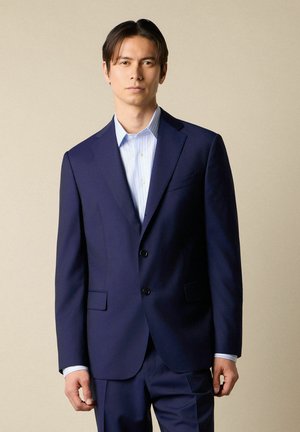 Man die een marineblauw pak en lichtblauw gestreept overhemd draagt, staand tegen een effen beige achtergrond, vooruitkijkend.