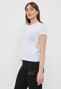 T-shirt bianco in cotone con maniche corte, con accentuazioni nere sulle spalle e collo rotondo. La vestibilità è casual e standard.