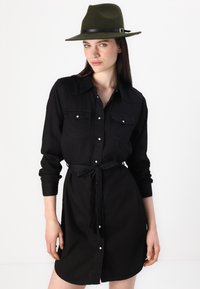 Rochie neagră din bumbac, cu deschidere cu nasturi, două buzunare pe piept, mâneci lungi, talie cu brâu și textură netedă. Asortată cu o pălărie din fetru verde.