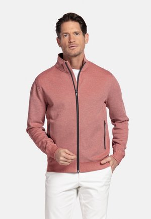 Man draagt een dusty rose jas met ritssluiting over een wit shirt en witte broek, staat met één hand in de zak tegen een effen achtergrond.