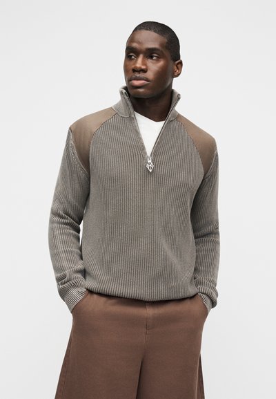 Άνδρας που φοράει καφέ πλεκτό πουλόβερ με φερμουάρ και καφέ παντελόνι, με σουέντ λεπτομέρειες στους ώμους, στέκεται με τα χέρια στις τσέπες σε λευκό φόντο.