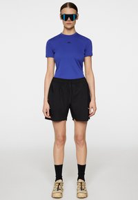 Blå åtsittande kortärmad skjorta, svarta shorts med elastiskt midjeband, svarta knähöga strumpor, beige skor med flerfärgade accenter, stora solglasögon.