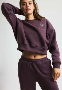 Felpa corta viola con scollo rotondo, polsini a costine e un piccolo logo. Abbinata a pantaloni jogger con vita elastica coordinati.