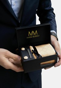 Una scatola regalo nera contenente un set di gemelli dorati, un fazzoletto da taschino a motivo dorato e una penna argentata, presentata da una persona in giacca e cravatta.