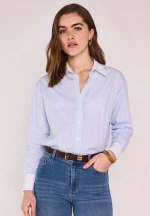 Frau trägt ein hellblau gestreiftes Hemd mit Knöpfen, das in eine hoch taillierte Jeans mit braunem Gürtel gesteckt ist, goldene Creolen und Armbänder.