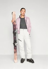 Roze geïsoleerd jasje over een grijs shirt, witte skibroek en zwarte skischoenen. Houdt zwarte en roze ski's met bindingen vast.