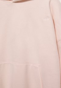 Sweat à capuche rose clair en tissu doux avec une poche kangourou et un logo brodé discret sur la poitrine. Texture lisse et coupe décontractée.
