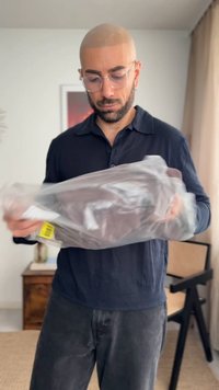 Άνδρας με γυαλιά και γενειάδα εξετάζει συσκευασμένα ρούχα μέσα σε ένα δωμάτιο με μια καρέκλα και μια κορνιζαρισμένη εικόνα στο φόντο.