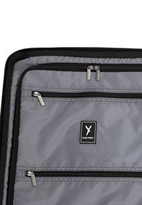 Grijze reistasseninterieur met twee zwarte ritscompartimenten. Voorzien van zilveren hardware en een merklabel met "SURI FREY." Glad textuur.