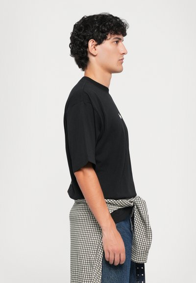 Giovane con capelli neri ricci che sta di lato, indossa una maglietta nera, jeans blu e una camicia a scacchi legata intorno alla vita.