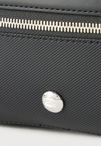 Κοντινό πλάνο μαύρης τσάντας με υφή, ασημένιο φερμουάρ και στρογγυλό μεταλλικό λογότυπο με έμβλημα κροκοδείλου στο μπροστινό πάνελ.