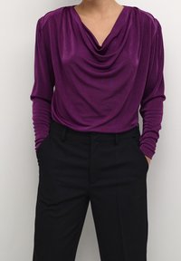 Haut col bénitier violet en tissu doux, à manches longues et design drapé, associé à un pantalon noir sur mesure.