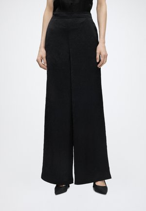 Femme portant un pantalon noir texturé, taille haute et jambes larges, avec des escarpins noirs pointus sur un fond blanc uni.
