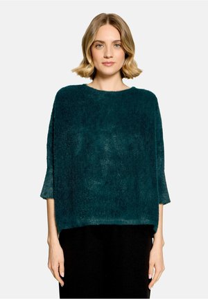 Vrouw met kort blond haar, die een donkergroene zachte trui met driekwartmouwen en een zwarte broek draagt, staand tegen een witte achtergrond.