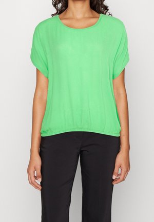 Kvinde iført en lysegrøn kortærmet bluse med elastisk kant og sorte bukser, stående foran en ensfarvet lys baggrund.