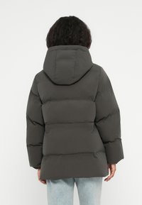 Mujer de espaldas vistiendo una chaqueta acolchada gris oscura con capucha y jeans azul claros contra un fondo blanco liso.