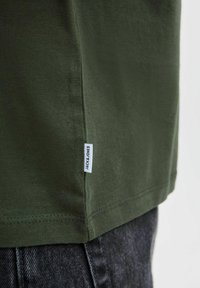Camiseta de algodón verde oliva con una etiqueta en la costura lateral. Textura suave, escote redondo y mangas cortas. El dobladillo termina a la altura de la cintura.