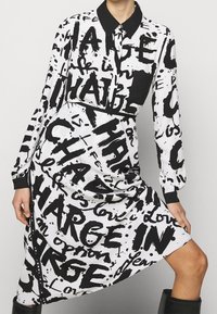 Abito a maniche lunghe con stampa testuale astratta in bianco e nero, dotato di colletto classico e polsini neri, con una vestibilità comoda e design fino al ginocchio.