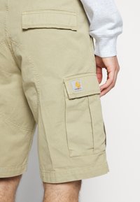 Short cargo beige avec poches à rabat et patch logo Carhartt sur la poche latérale, porté par une personne portant un pull gris à manches longues.