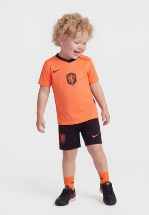 Enfant aux cheveux bouclés souriant, portant un maillot de football orange et noir avec un blason de lion, des chaussettes orange et des crampons Nike noirs.