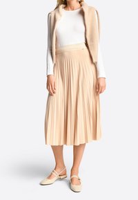 Falda beige plisada con una cinturilla texturizada, combinada con una blusa de manga larga clara y unos zapatos planos beige con un diseño de tiras.