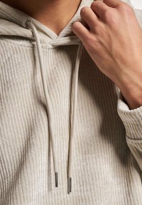 Beige ribbestrikket hettegenser med snøring. Har glatte, blanke detaljer og et teksturert stoff. Hånd som griper snoren er synlig.