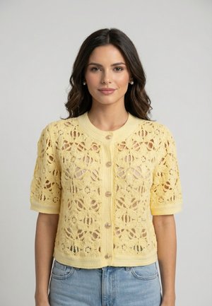 Donna con capelli scuri e mossi indossa una maglietta gialla all'uncinetto con bottoni e maniche corte e jeans azzurri chiari su uno sfondo grigio semplice.