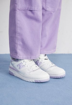 Zapatillas blancas y lavanda combinadas con pantalones amplios de color púrpura claro, de pie sobre un suelo alfombrado de azul.